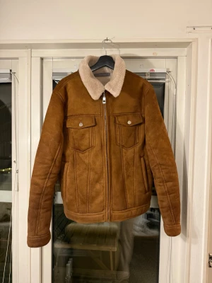 Brun teddyjacka från Zara Man - Säljer en brun teddyjacka från Zara Man med mjuk, beige fuskpäls på kragen. Jackan har två bröstfickor med lock och knappar, samt dragkedja framtill. Jackan har en klassisk, rak passform.