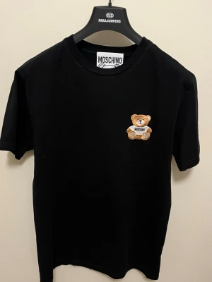Svart Moschino t-shirt med nalle - Svart t-shirt från Moschino med en broderad nalle på bröstet. Klassisk rund halsringning och korta ärmar. T-shirten har en stilren och trendig look som passar perfekt till jeans eller mjukis 