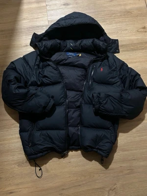 Svart dunjacka från Polo Ralph Lauren - Snygg svart dunjacka från Polo Ralph Lauren med röd logga på bröstet. Jackan har huva, dragkedja framtill och flera fickor. Puffig modell som håller dig varm under kalla dagar. Perfekt för dig som vill ha både stil och funktion.