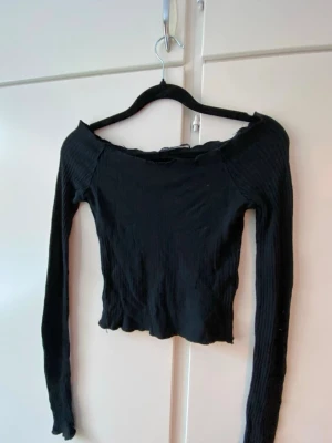 Svart ribbad off shoulder topp - Trendig svart långärmad topp med ribbad struktur och off shoulder-design. Croppad modell med vågig kant nertill och vid ärmslut. Perfekt att styla med höga jeans eller kjol för en cool look.