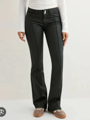 Svarta bootcut jeans med dubbla knappar - Snygga svarta bootcut jeans med dubbla knappar i midjan och markerade sömmar längs benen. Jeansen har en hög midja och är tillverkade i ett glansigt material som ger en cool look. Perfekta för dig som vill sticka ut lite extra.