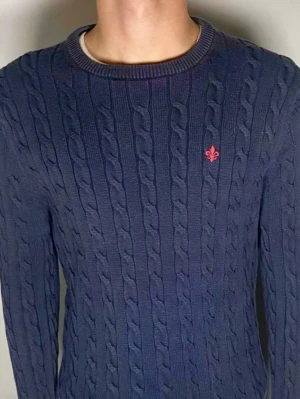 Mörkblå kabelstickad tröja St L - Mörkblå kabelstickad tröja med rund halsringning och ett rött broderat emblem på bröstet. Tröjan har långa ärmar och ribbade muddar vid ärmslut och nederkant. Perfekt för dig som gillar klassisk och stilren design. Storlek Large. Snabb Leverans!