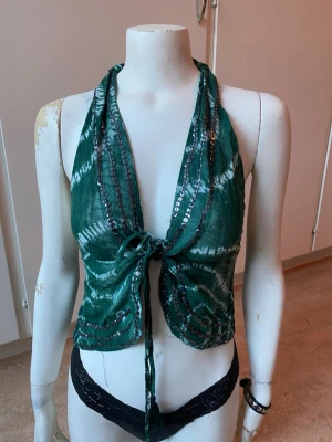Grön halterneck topp med paljetter - Snygg grön halterneck topp med knyt framtill och öppen rygg. Toppen har ett batikinspirerat mönster och är dekorerad med paljetter längs kanterna. Perfekt för festival eller bara soliga dagar! 