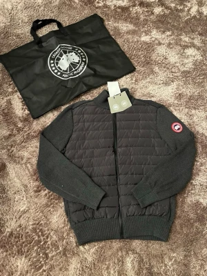 Svart cardigan jacka från Canada Goose - Stilren svart cardigan jacka från Canada Goose med quiltad front och stickade ärmar samt rygg. Jackan har dragkedja framtill och klassisk logotyp på vänster ärm. Perfekt för lager-på-lager och snygg till många outfits.