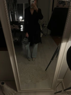 Svart omlottkappa med bälte - Stilren svart kappa i omlottmodell med knytbälte i midjan. Kappan har en klassisk krage och lång ärm, vilket ger en elegant och tidlös look. Perfekt att slänga över en outfit för en chic vibe. Mycket bra skick