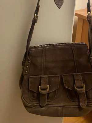 Brun axelväska i skinn med spännen - Snygg brun axelväska i skinn med justerbar rem och två framfickor med spännen. Väskan har dragkedja upptill och detaljer i mässing. Perfekt för dig som vill ha en stilren och praktisk väska med coola detaljer.