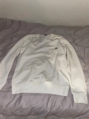Vit sweatshirt från Lacoste - Säljer en stilren vit sweatshirt från Lacoste med klassisk rund halsringning och det ikoniska krokodilmärket broderat på bröstet. Tröjan har långa ärmar och ribbade muddar vid ärmslut och nederkant. Perfekt för en clean och avslappnad look.