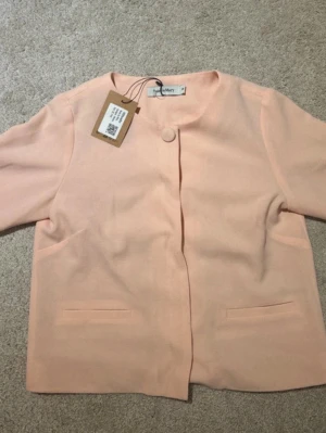 Ljusrosa kavaj från Rules by Mary - Stilren ljusrosa kavaj från Mary med rund halsringning och enkel knappstängning framtill. Kavajen har raka, långa ärmar och två diskreta fickor framtill. Perfekt för en clean och modern look.