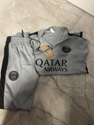 Grå PSG träningsset - Snyggt grått träningsset från PSG med svarta detaljer och klubbens logga på både tröja och byxor. Setet har dragkedja i halsen och elastisk midja på byxorna. Materialet är troligtvis polyester, perfekt för träning eller chill