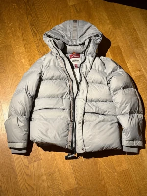 Parajumper jacka - Snygg grå dunjacka från parajumper med huva och dragkedja framtill. Jackan har en klassisk pufferstil och breda kanaler, perfekt för kalla dagar. Fodrad och med ribbade muddar vid ärmslut. Passar dig som vill ha både stil och värme. Skulle säga att den passar runt 160-165, ifall du har några frågor skriv. Pris är ej hugget i sten