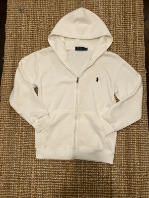Vit hoodie från Polo Ralph Lauren - Klassisk vit hoodie från Polo Ralph Lauren med dragkedja framtill och huva. Har två fickor och den ikoniska lilla svarta loggan broderad på bröstet. Perfekt för en clean och stilren look. Mjuk bomullskänsla och ribbade muddar. Jag har sänkt priset på den eftersom snörena försvann i tvättmaskinen 