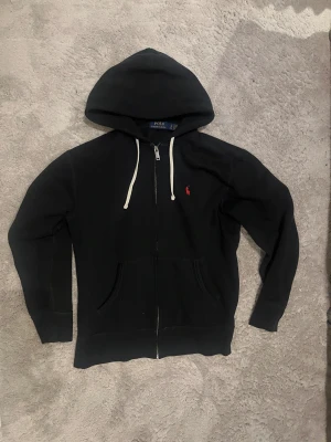 Svart hoodie från Polo Ralph Lauren - Svart hoodie med dragkedja från Polo Ralph Lauren. Klassisk design med vit dragsko i huvan och röd broderad logga på bröstet. Bra skick och riktigt bra kvalitet!