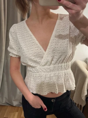 Vit omlottopp med broderade detaljer - Superfin vit omlottopp med broderade hål och volang nertill. Toppen har korta puffärmar, markerad midja och v-ringning. Perfekt att styla med jeans eller kjol för en trendig look.