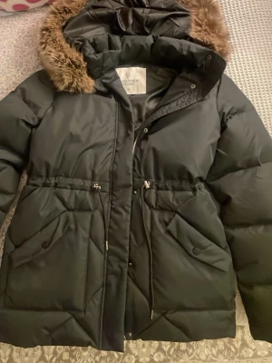 Svart dunjacka med fuskpäls från Bubbleroom - Svart dunjacka från Bubbleroom med huva och avtagbar fuskpäls. Jackan har dragsko i midjan, två stora fickor framtill och stängs med dragkedja och knappar. Perfekt för kalla dagar med sin varma och fluffiga design.