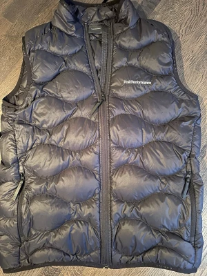 Svart dunväst från Peak Performance - Snygg svart dunväst från Peak Performance med hög krage och två dragkedjefickor. Västen har en quiltad design och Peak Performance-logga på bröstet. Perfekt för lager-på-lager och håller dig varm under kyliga dagar.