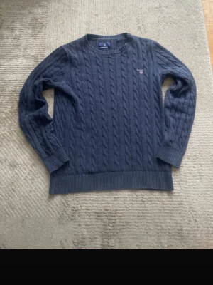 Mörkblå kabelstickad tröja från GANT - Kabelstickad mörkblå tröja från GANT med rund halsringning och ribbade muddar. Tröjan har GANT-logga broderad på bröstet och är tillverkad i ett mjukt, tjockt material som passar perfekt för kyligare dagar. Klassisk och stilren design.