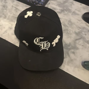 Svart keps från Chrome Hearts - Säljer en svart keps från Chrome Hearts med vita broderade kors och CH-logga framtill. Kepsen har böjd skärm och detaljer med Chrome Hearts-tryck på insidan. Snygg och streetinspirerad design med metallknapp på toppen.