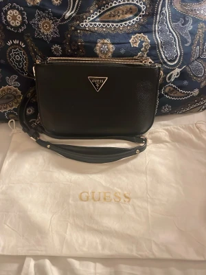 Svart axelväska från Guess - Snygg svart axelväska från Guess med justerbar axelrem och guldfärgade detaljer. Väskan har ett klassiskt trekantigt Guess-emblem framtill och stängs med dragkedja. Perfekt storlek för det viktigaste och passar till många olika outfits.OBS!! Det har kommit en vitt fläck på väskan(se bild nmr3). Den syns inte särskilt mycket. Köpt för 1199kr på room 1006 för ett år sedan.