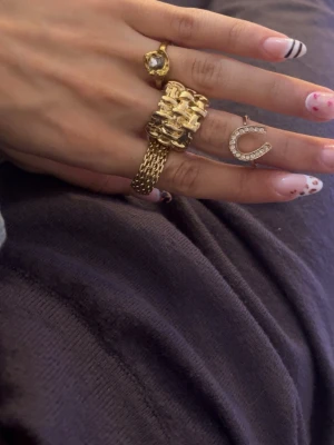 Guldiga ringar med stenar och kedjedetaljer - Fyra unika ringar i guldton: en med stor klar sten, en bred med kedjedetaljer och en tunnare med hästskoformad dekoration täckt av små glittrande stenar. Perfekta för att skapa en trendig och personlig stil. 40kr styck