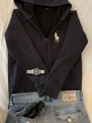 Svart hoodie från Polo Ralph Lauren - Säljer en svart hoodie från Polo Ralph Lauren med dragkedja och stor vit logga på bröstet. Klassisk huva och två fickor framtill. Tillverkad i mjukt bomullsmaterial som är skönt att ha på sig. Perfekt för en avslappnad och stilren look. Köpte på nån marknad. Sitter bra och tycker om den