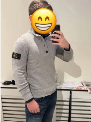 Grå stickad tröja från Stone Island - Snygg grå stickad tröja från Stone Island med hög krage och knappar framtill. Tröjan har lång ärm, ribbade muddar och den klassiska logotypen på vänster ärm. Perfekt för dig som gillar streetwear och vill ha något stilrent och bekvämt.
