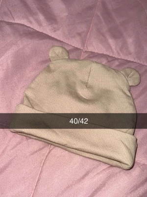 Beige ribbad mössa med björnöron - Supersöt beige mössa i ribbad bomull med vikbar kant och små björnöron på toppen. Perfekt accessoar för dig som vill ha en lekfull och trendig look. Storlek 40/42.