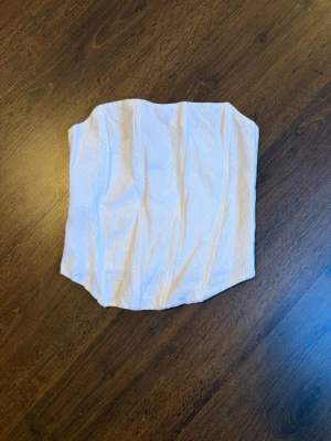 Vit bandeau bustier med broderi - Säljer en vit bandeau bustier med diskret blommigt broderi. Toppen har en figurnära passform och smock i ryggen för extra komfort. Perfekt att styla under en skjorta eller som den är till höga jeans eller kjol.
