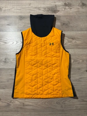 Under Armour väst - Sportig gul väst från Under Armour med svart huva och svarta detaljer längs kanterna. Västen har ett quiltat mönster framtill och är ärmlös, perfekt för lager-på-lager. Under Armour-logga på bröstet ger en clean och modern look.