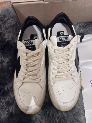 Golden Goose skor  - Snygga vita sneakers från Golden Goose med svart stjärndetalj på sidan och svarta detaljer vid hälen. Skorna har snörning, perforerad tå och platt sula. Tillverkade i skinn och har en klassisk, sportig look.