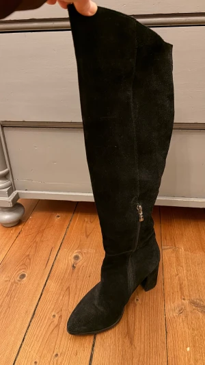 Svarta höga mockastövlar med klack - Säljer ett par svarta, höga stövlar i mjuk mocka med spetsig tå och stabil klack. Stövlarna har dragkedja på sidan för enkel påtagning och en stilren, minimalistisk look som passar till många outfits.