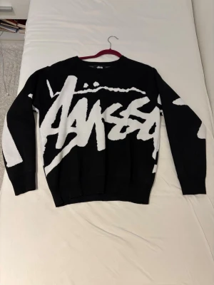 Svart och vit stickad tröja Stüssy - Säljer en svart stickad tröja från Stüssy med stort vitt grafiskt logotyptryck över hela framsidan och ärmarna. Tröjan har rund halsringning och ribbade muddar. Perfekt för dig som gillar streetwear och vill sticka ut.