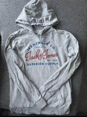 Grå hoodie från Jack & Jones - Ljusgrå hoodie från Jack & Jones med tryck i rött och blått framtill. Pris kan diskuteras vid snabb affär 