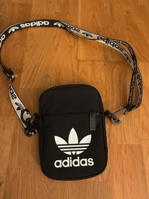 Svart axelväska från Adidas - Snygg svart axelväska från Adidas med vit logga framtill och justerbart axelband med Adidas-tryck. Väskan har dragkedja och är tillverkad i slitstark polyester. Perfekt för att bära det viktigaste och ge din outfit en sportig touch.