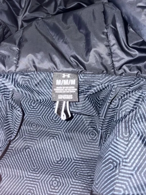 Svart dunjacka från Under Armour - Snygg svart dunjacka från Under Armour med quiltad design och dragkedja framtill. Jackan har en hög krage och diskret logga på bröstet. Perfekt för kalla dagar och har ett modernt, sportigt uttryck. Denna jacka är lite längre ner till rumpan. Nypris 2500kr