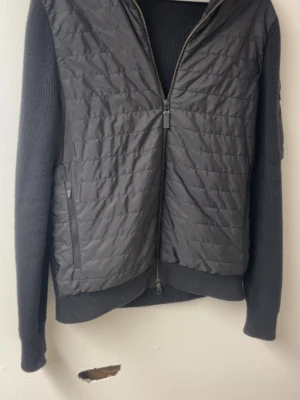 Svart overshirt från J.Lindeberg - Stilren svart overshirt från J.Lindeberg med quiltad front och stickade ärmar. Jackan har dragkedja framtill, diskret logga på ärmen och sidofickor med dragkedja. Perfekt lager-på-lager-plagg för en modern look.