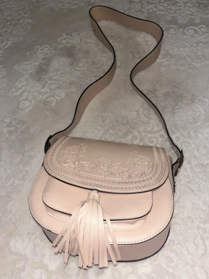 Beige axelväska med tofs och blommor - Snygg beige axelväska i skinnimitation med blommigt mönster på locket och en stor tofs framtill. Väskan har rundad form och justerbar axelrem. Perfekt för dig som vill ha en stilren men ändå unik accessoar. Bandet på väskan är bara lite smutsig på just den delen av bilden jag tog.