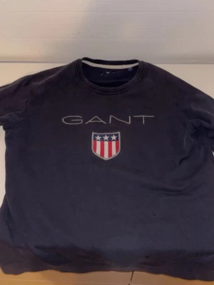 Mörkblå sweatshirt från GANT - Säljer en mörkblå sweatshirt från GANT med klassisk logga och sköld med stjärnor och ränder på bröstet. Tröjan har rund halsringning och mjukt material, perfekt för en avslappnad stil.