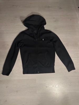 Svart zip hoodie från Polo Ralph Lauren - Snygg svart hoodie med dragkedja från Polo Ralph Lauren. Klassisk design med huva och dragsko, två fickor fram och den ikoniska lilla loggan broderad på bröstet. Tillverkad i mjukt bomullsmaterial som är skönt att bära.