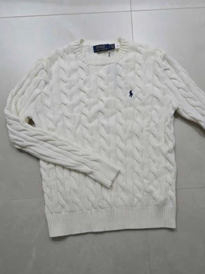 Vit kabelstickad tröja från Polo Ralph Lauren - Kabelstickad vit tröja från Polo Ralph Lauren med rund halsringning och blå broderad logga på bröstet. Tröjan har långa ärmar och ribbade muddar vid ärmslut och nederkant. Perfekt för en klassisk och stilren look.