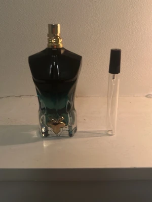 Le Male parfym från Jean Paul Gaultier - Ikonisk parfymflaska formad som en manlig torso i glas med mörkblå och gröna toner samt gulddetaljer på korken. Medföljer en genomskinlig provflaska med svart lock. Perfekt för dig som gillar unika och snygga flaskor.