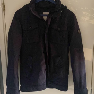 Svart vindjacka från J.Lindeberg - Säljer en svart vindjacka från J.Lindeberg i storlek M. Jackan har huva, fyra fickor med lock och knappar samt dragkedja framtill. Snygg metallbadge på ärmen och klassisk, stilren design. Perfekt för dig som vill ha en enkel men trendig jacka.