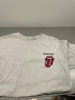Rolling Stones vit t-shirt - Vit t-shirt med Rolling Stones klassiska logga framtill och stor svart text på ryggen. Rund halsringning och korta ärmar. Perfekt för dig som gillar bandtröjor och vill sticka ut med en ikonisk design.
