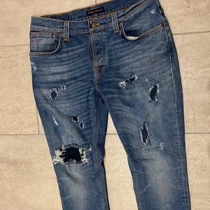Blå slitna jeans från Nudie Jeans - Säljer ett par blå jeans från Nudie Jeans i storlek 34/32. De har flera slitningar och hål på framsidan för en rå och edgy look. Klassisk femficksmodell med orange sömmar och broderade bakfickor. Perfekt för dig som gillar streetstyle.