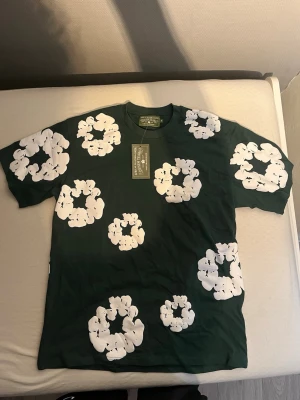 Grön t-shirt med vita blommor Denim Tears - Snygg grön t-shirt från Denim Tears med stora vita blommor tryckta över hela tröjan. Klassisk rund hals och korta ärmar.                                                              Strlk M men passar L