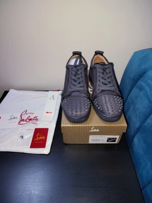 Svarta sneakers från Christian Louboutin - Säljer ett par svarta sneakers från Christian Louboutin med rund tå och coola nitar på tån. Skorna har snörning och är tillverkade i ett tygmaterial med läderdetaljer. Perfekta för dig som vill sticka ut med en edgy look.