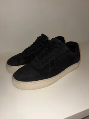 Svarta sneakers från Axel Arigato - Stilrena svarta sneakers från Axel Arigato med ovandel i mocka och klassisk vit sula. Skorna har rund tå, snörning och diskret logga i guld på sidan. Perfekta för dig som gillar minimalistisk design och vill ha ett par sneakers som funkar till allt.