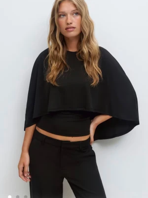 Svart croppad bolerotopp - Stilren svart croppad bolerotopp med vid, cape-liknande överdel. Toppen har en modern och edgy look, perfekt för att sticka ut. Materialet känns mjukt och följsamt mot kroppen. Passformen är normal och toppen är ärmlös under den vida bolerodelen.