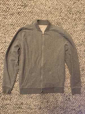 Rose and born zip - Snygg grå zip från rose and born med minimalistisk design och ribbade muddar. Tröjan har dragkedja framtill, två sidofickor och klassisk bomberkrage. Perfekt för dig som gillar enkel och clean stil. Taggen vid nacken har fallit av