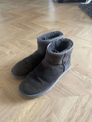 Gråa UGGS skor i mocka - Mysiga gråa tofflor från UGG i mjuk mocka med fluffigt foder. Modellen har en högre ankel och rund tå, perfekt för att hålla fötterna varma hemma. Klassisk UGG-logga bak på hälen och platt sula för extra komfort.