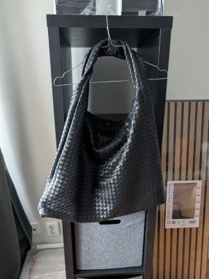 Svart flätad handväska i syntet - Stilren svart handväska med flätad design och mjuk form. Väskan har bred axelrem och stängs med knapp. Tillverkad i syntetmaterial som ger en lyxig känsla. Perfekt för dig som vill ha en trendig och rymlig väska till vardagen.